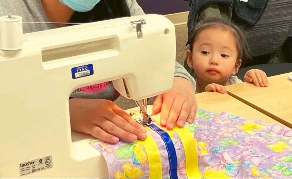 A person sewing ribbon onto fabric with a sewing machine while a small child watches